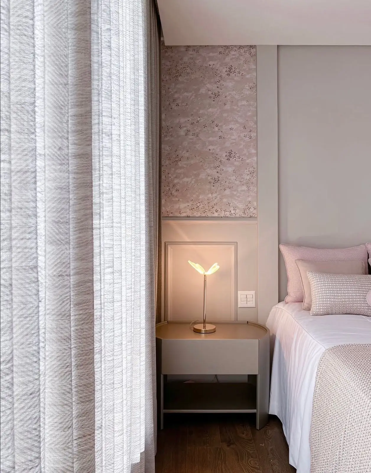 Quarto com cortina clara, papel de parede rosado e luminária de cabeceira