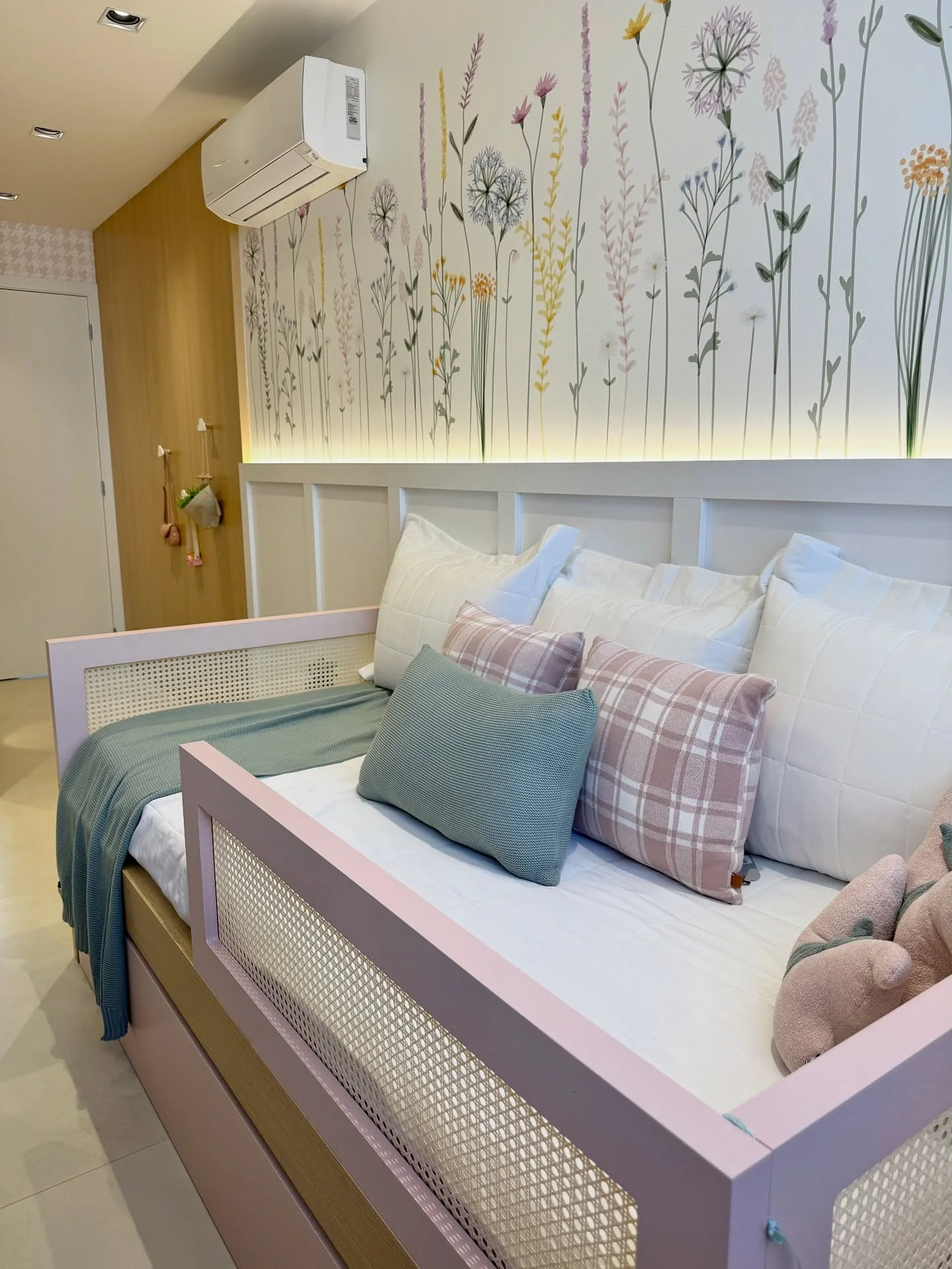 Cama rosa e madeira com almofadas, sob parede floral e iluminação indireta