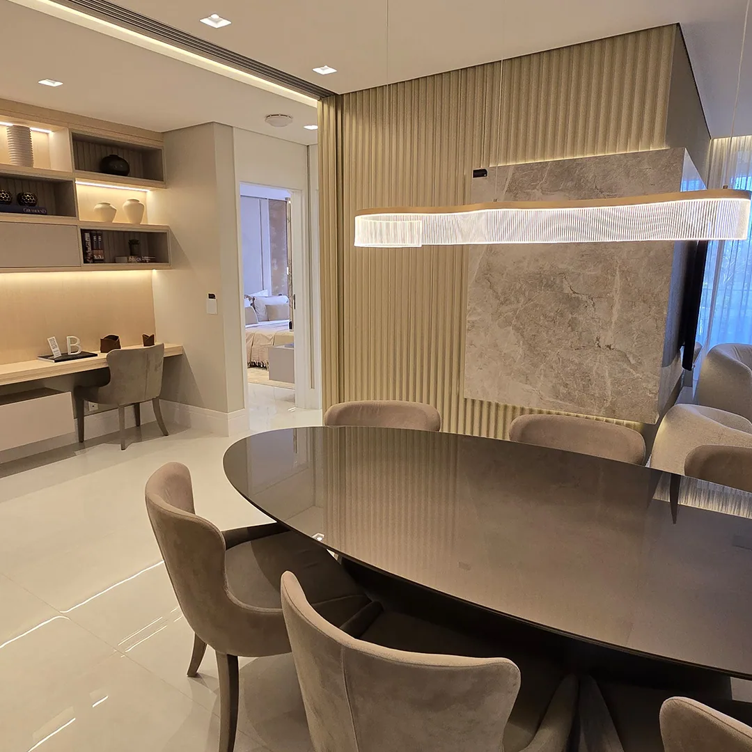 Sala de jantar com mesa oval escura, cadeiras estofadas, pendente em anel e painel vertical atrás da televisão