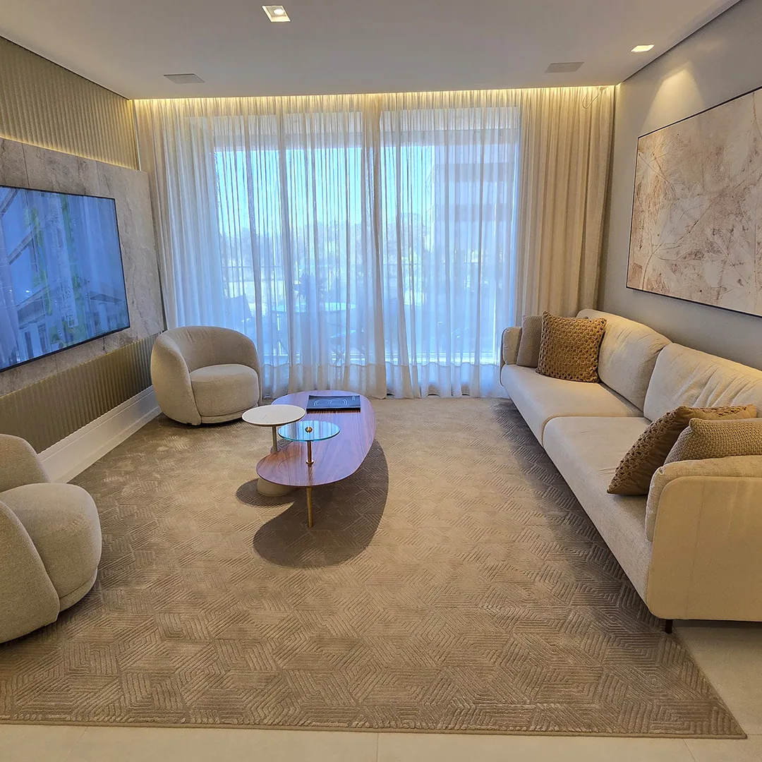 Sala com sofá claro, duas poltronas arredondadas, tapete geométrico e cortina translúcida do piso ao teto