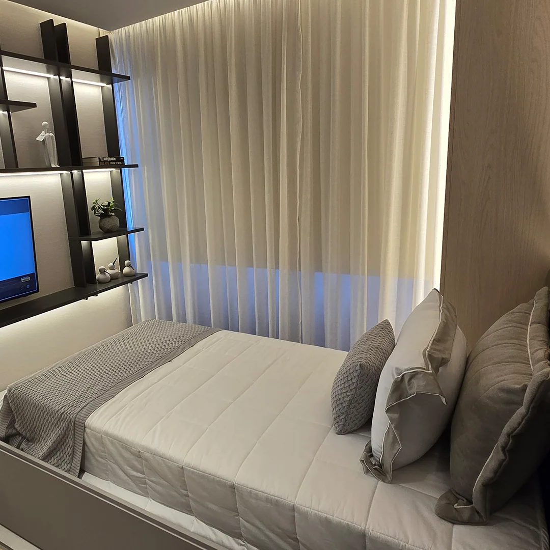 Quarto com cama clara, cabeceira estofada, cortina do piso ao teto e televisão ao lado de prateleiras escuras iluminadas