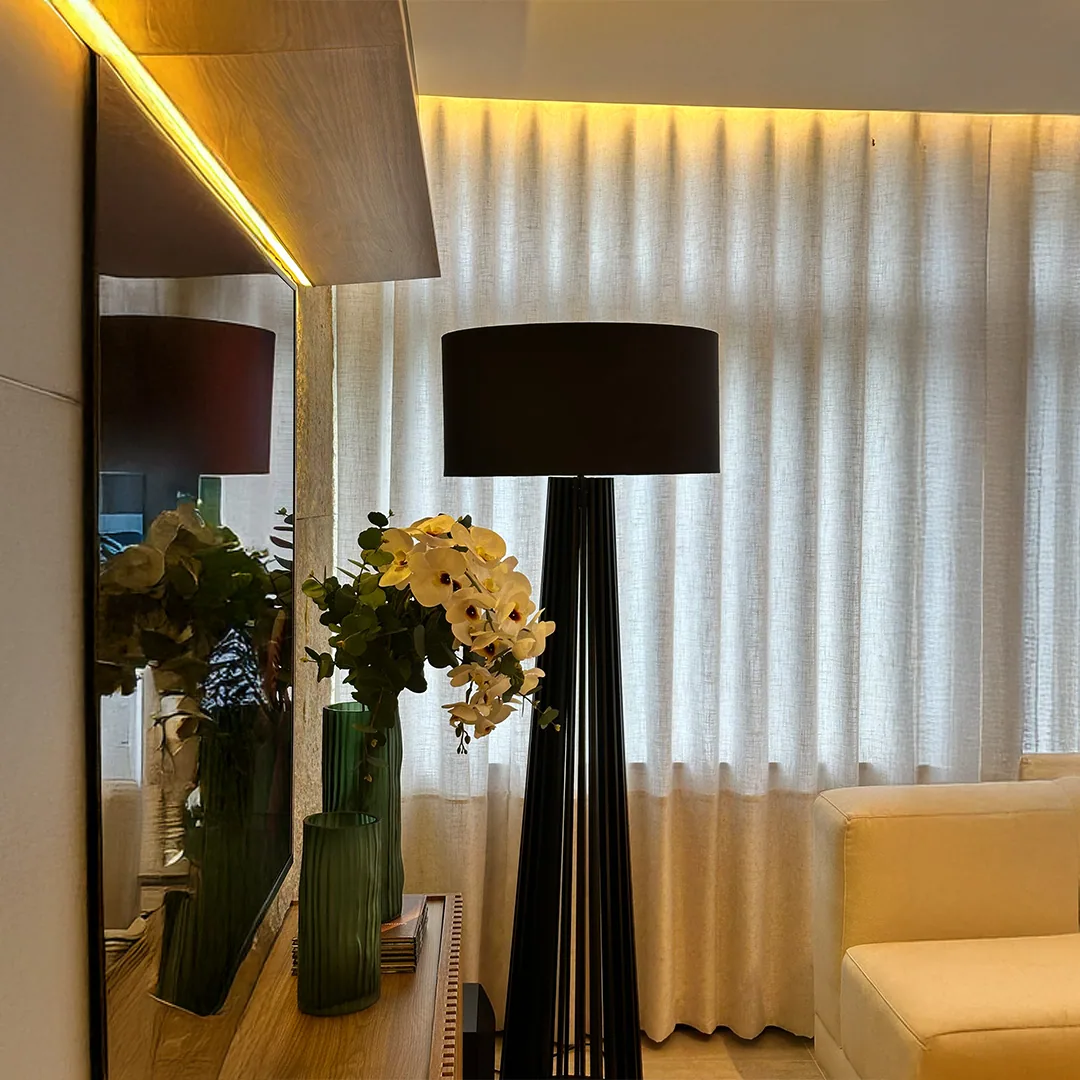 Detalhe de sala com cortina clara, espelho vertical, iluminação linear, luminária preta, sofá claro e vasos verdes