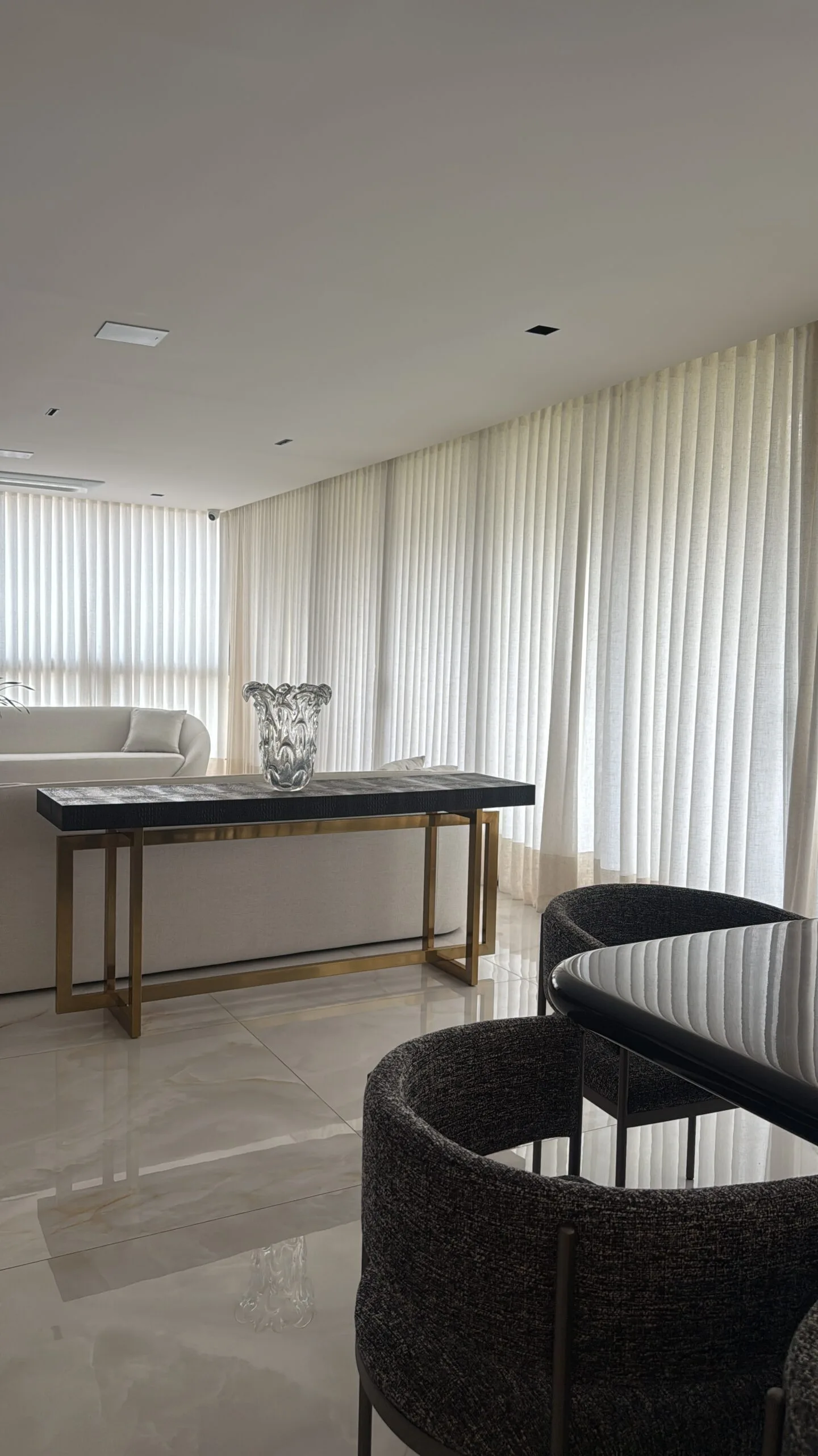 Cortinas claras com dobras verticais junto a uma sala com sofás claros e aparador escuro