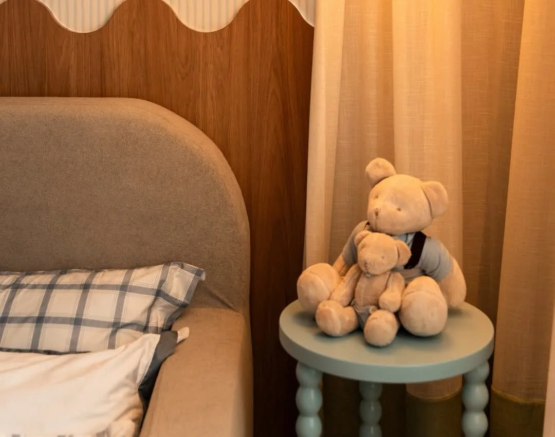 Cama clara, mesa lateral azul com ursinhos, cabeceira ondulada e cortina no quarto infantil de Fernanda Lettieri