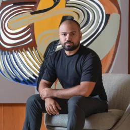 Raphaell Cruz sentado diante de uma obra de formas coloridas